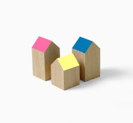 Drie houten blokjes in de vorm van een huisje met schuine daken gekleurd in roze, blauw en geel